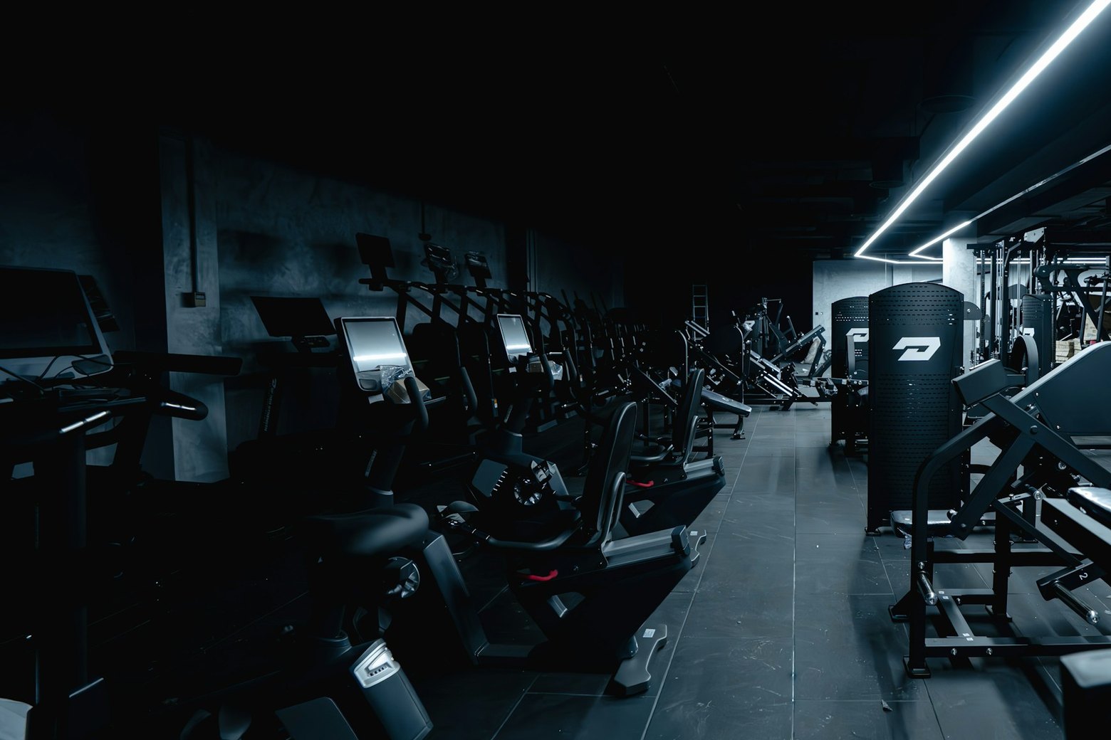 Pasillo de máquinas en un gimnasio vacío de noche, iluminado por una sola franja de luz en el techo